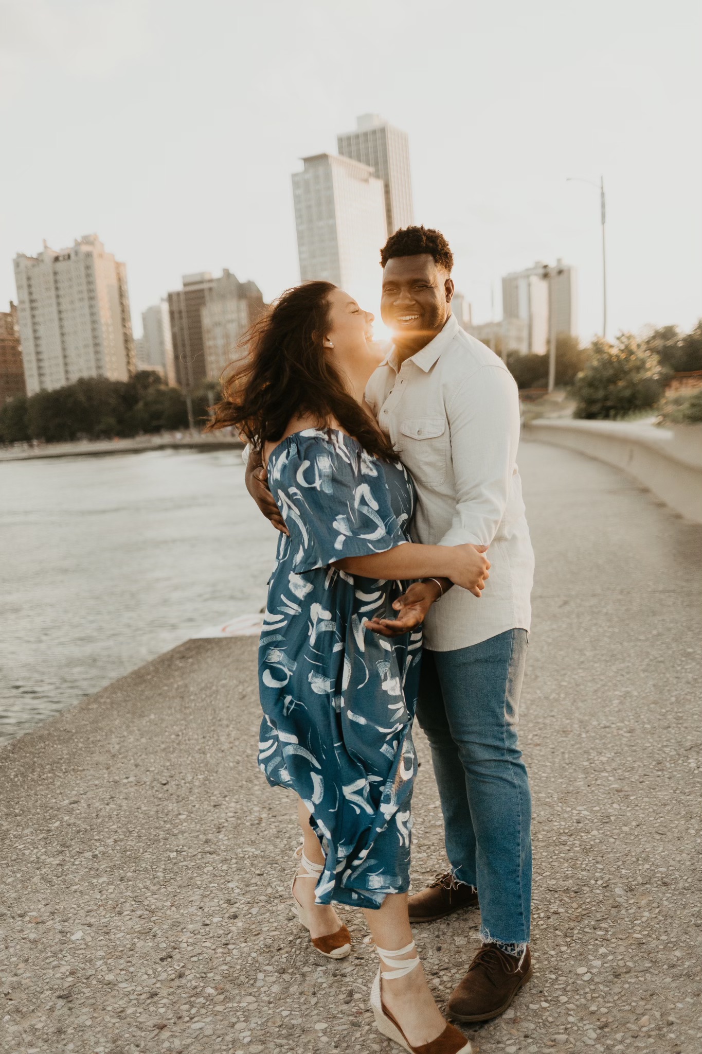 A joyful couple embracing near the water on a scenic Chicago lakeside path, with the city skyline in the background. The woman wears a flowing blue dress with abstract white patterns, while the man wears a light-colored button-up shirt and jeans. The sunlight casts a warm glow, highlighting their smiles.