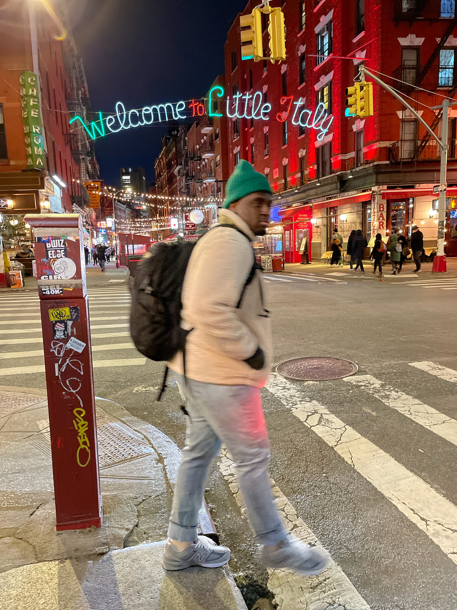 Man in a green beanie and beige fleece crossing the street in Little Italy, New York City at night, with the 'Welcome to Little Italy' neon sign glowing in the background.