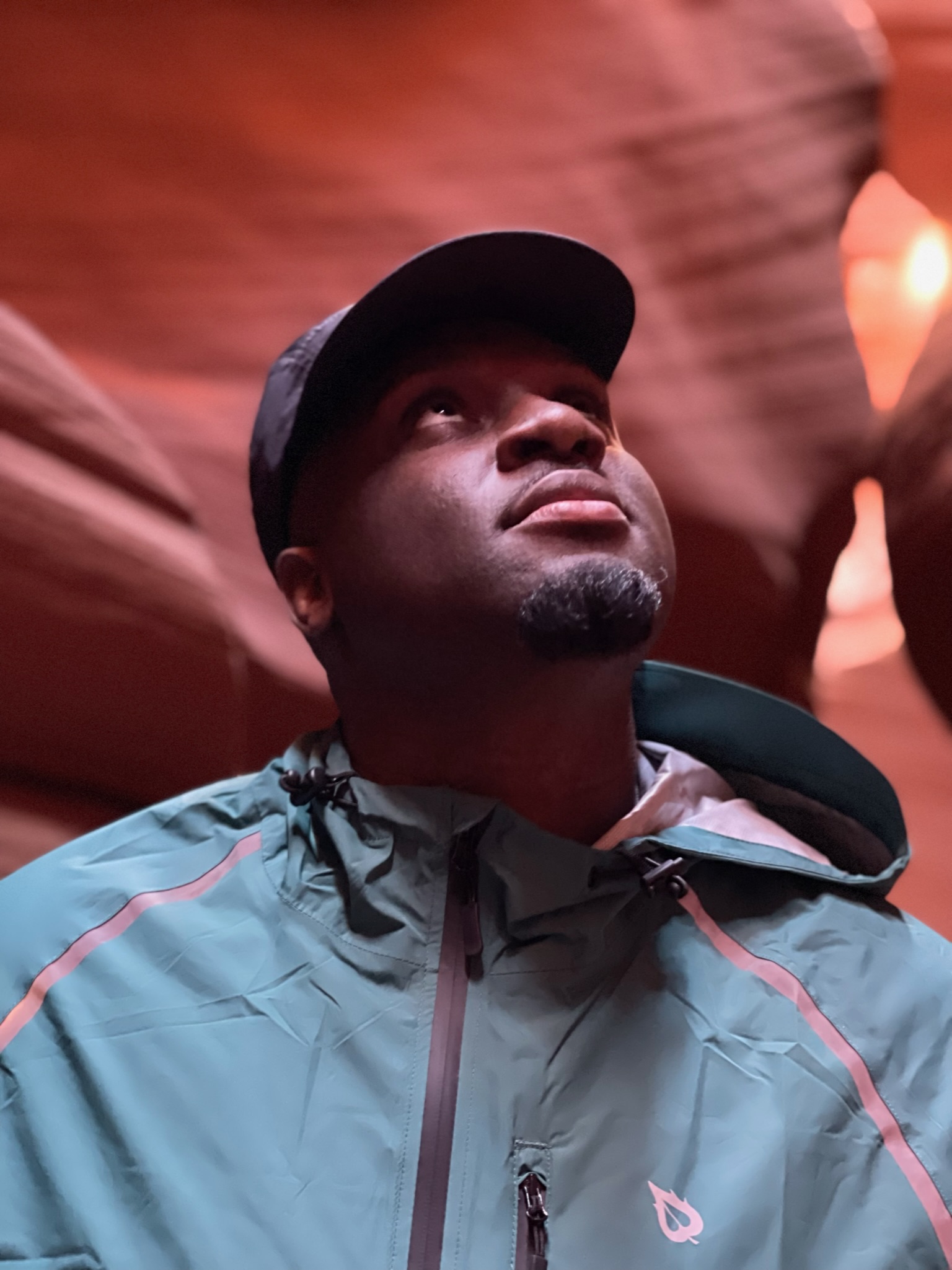 Man in a teal jacket and black cap gazing upward in Antelope Canyon, Arizona. The red sandstone walls curve around him, illuminated by soft natural light.